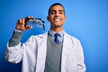 Yakışıklı afro-amerikan optik adam mavi arka planda optometri gözlüğü tutuyor mutlu bir yüzü var ve gülümsüyor kendine güvenen bir gülümsemeyle dişlerini gösteriyor