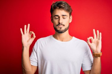 Kırmızı arka planda dikilen sıradan tişörtlü, sakallı, genç ve yakışıklı bir adam. Gözleri kapalı, meditasyon yaparken parmaklarıyla gülümsüyor. Yoga konsepti.