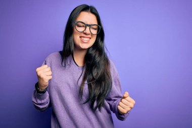 Mor izole edilmiş arka planda gözlük takan genç esmer kadın mutlu ve heyecanlı bir şekilde kollarını kaldırıp, gülümseyerek ve başarı için çığlık atarak kazanan bir jest yapıyor. Kutlama konsepti.