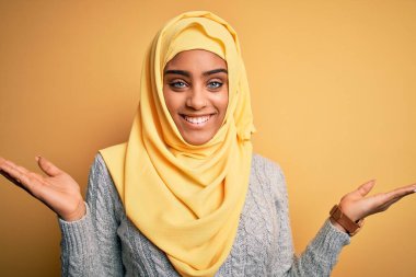 Genç ve güzel Afro-Amerikalı kız izole edilmiş sarı arka planda Müslüman başörtüsü takıyor gülümsüyor iki elini de açık avuçlarında gösteriyor, sunuyor ve reklam karşılaştırması yapıyor ve dengeyi kuruyor.