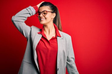 Güzel, esmer, ceketli, kırmızı arka planda gözlüklü, kendine güvenen, el hareketleriyle saçlarına dokunan çekici ve modaya uygun bir iş kadını.