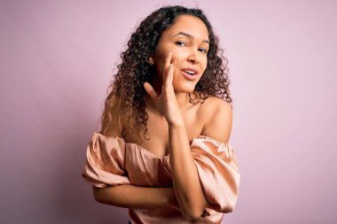 Kıvırcık saçlı, pembe tişörtlü genç bir kadın pembe arka planda duruyor. Ağzında gizli dedikodular dolaşıyor, kötü niyetli konuşmalar yapıyor.