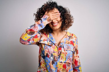 Genç ve güzel kıvırcık Arap kadın çiçek rengi gömlek giyiyor beyaz arka planda duruyor gülümsüyor ve elleri yüzünde şaşkınlık için gözleri kapatıyordu. Kör kavram.