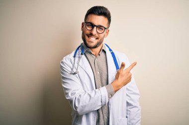 Gözlük takan genç doktor, tıbbi beyaz cübbe ve steteskop ile izole edilmiş arka planda neşeli, yüzünde mutlu ve doğal bir ifade olan el ve parmak ile gülümseyen bir yüz.