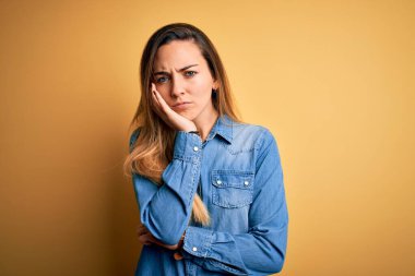 Sarı arka planda kot gömlek giyen, mavi gözlü genç ve güzel sarışın kadın kollarını kavuşturarak depresyondan sıkılmış ve yorgun görünüyor..