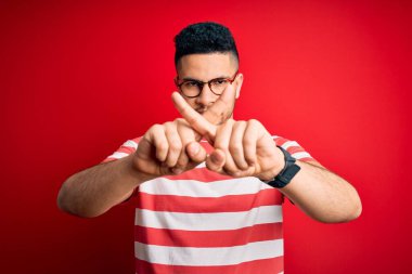 Genç ve yakışıklı bir adam günlük çizgili tişört ve gözlüklerle izole edilmiş kırmızı arkaplan üzerinde reddedilme ifadesini negatif işaret yaparak umut ediyor.