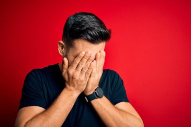 Siyah bir tişört giyen yakışıklı bir genç, izole edilmiş kırmızı arka planda dikiliyor. Üzücü bir yüz ifadesi var. Ağlarken yüzünü kapatıyor. Depresyon kavramı.