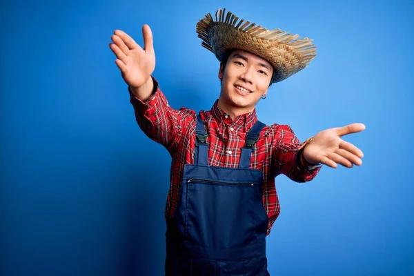 Young handsome chinese farmer man wearing apron and straw hat over blue background looking at the camera smiling with open arms for hug. Cheerful expression embracing happiness.