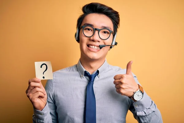 Young handsome chinese call center agent man holding paper with question mark symbol happy with big smile doing ok sign, thumb up with fingers, excellent sign