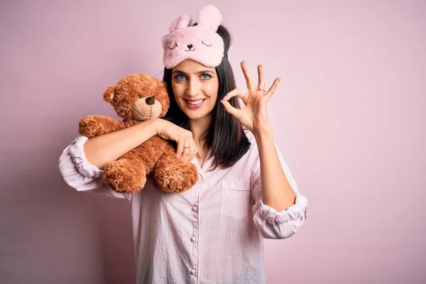 Young brunette woman with blue eyes wearing pajama hugging teddy bear stuffed animal doing ok sign with fingers, excellent symbol