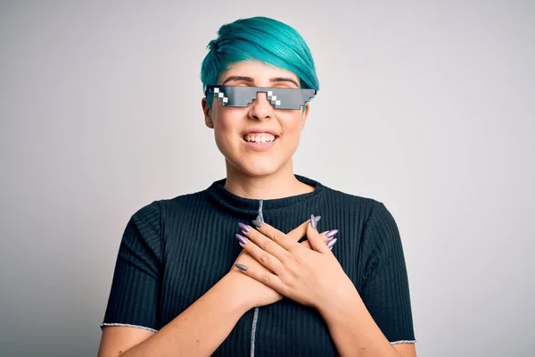 Young woman with blue fashion hair wearing thug life sunglasses over white background smiling with hands on chest with closed eyes and grateful gesture on face. Health concept.