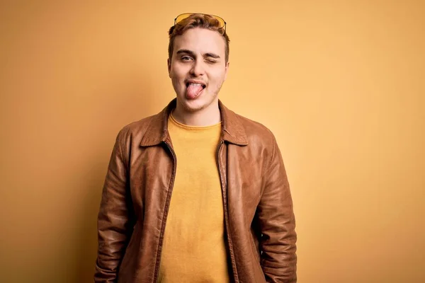 Young handsome redhead man wearing casual leather jacket over isolated yellow background sticking tongue out happy with funny expression. Emotion concept.