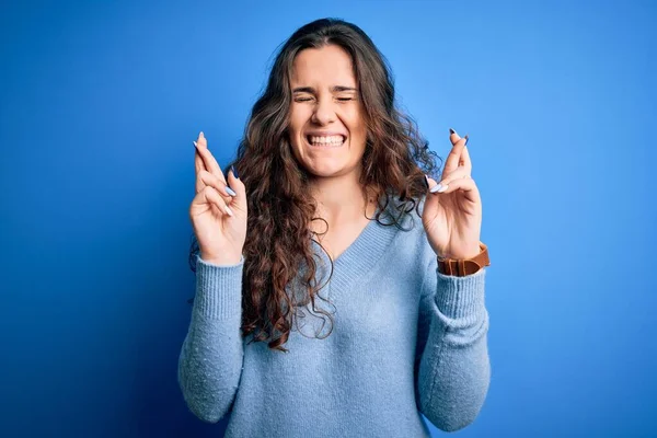 Young beautiful woman with curly hair wearing blue casual sweater over isolated background gesturing finger crossed smiling with hope and eyes closed. Luck and superstitious concept.