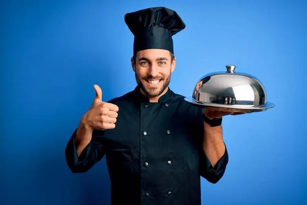 年轻的厨师 留着胡子 身穿制服 拿着圆顶蓝色背景的盘子 带着大大的笑容 做得不错 手指头翘起 是极好的标志 — 图库照片