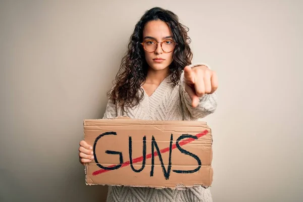 Young beautiful woman with curly hair holding banner with prohibited guns message pointing with finger to the camera and to you, hand sign, positive and confident gesture from the front