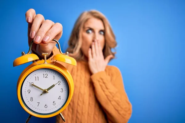 Middle age beautiful blonde woman holding vintage alarm clock over blue background cover mouth with hand shocked with shame for mistake, expression of fear, scared in silence, secret concept