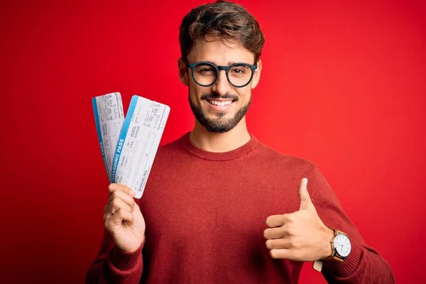 休暇中の若い観光客の男性搭乗券を保持する上で大きな笑顔で幸せな赤い背景の上に立って をサインを行うと 親指を指で開くと 優れたサイン — ストック写真