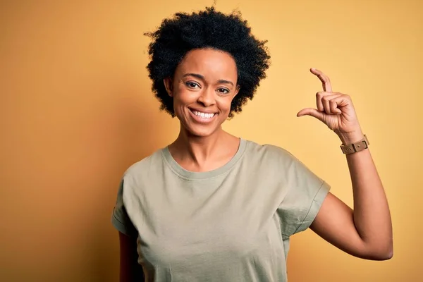 カジュアルなTシャツを着た巻き毛をした若い美しいアフリカ系アメリカ人アフロ女性は笑顔で 指を見てカメラで小さなサイズのサインをして手で自信を持ってジェスチャー 概念の測定 — ストック写真