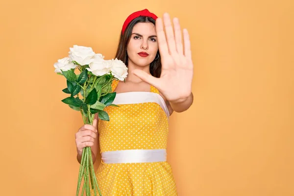 Young beautiful pin up woman wearing 50s fashion vintage dress holding boquet of roses flowers with open hand doing stop sign with serious and confident expression, defense gesture