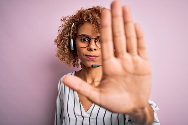 Африканский американец кудрявый агент колл-центра женщина работает с помощью гарнитуры на розовом фоне делать остановить петь с ладонью руки. Предупреждающее выражение с негативным и серьезным жестом на лице
.