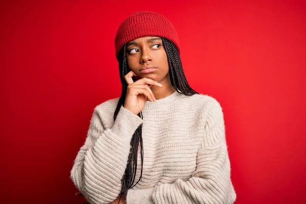 Young african american woman wearing winter sweater and wool hat over red isolated background with hand on chin thinking about question, pensive expression. Smiling with thoughtful face. Doubt concept.