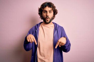 Genç, yakışıklı, sakallı, pembe arka planda sıradan bir kazak giyen, reklamını yapan, şaşırmış suratlı ve ağzı açık genç bir adam.