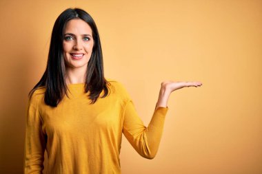 Mavi gözlü genç esmer kadın sarı arka planda günlük süveter giyiyor neşeli bir şekilde gülümsüyor ve avuç içi kameraya bakıyor..