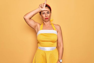 Orta yaşlı bir kadın, 50 'li yaşlarda, sarı arka planda eski tarz bir elbise giyiyor. Alnında parmakları olan insanlarla alay ediyor. Ezik jestler yapıyor ve aşağılıyor..