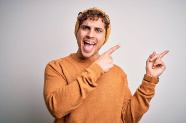 Sarışın, kıvırcık saçlı, günlük kazak giyen, yün şapkalı, gülümseyen ve kameraya bakan iki eli ve parmağı yana doğru bakan genç bir adam..