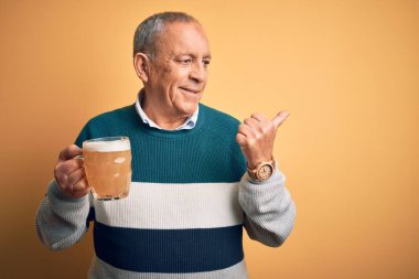 Üst düzey yakışıklı adam izole edilmiş sarı arka planda duran bira kavanozunu içerken gülümsüyor ve başparmağıyla yan tarafa bakıyor..