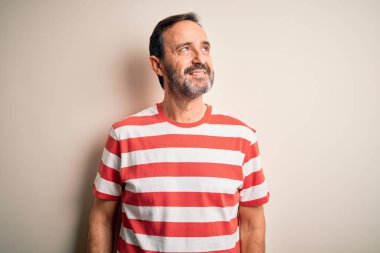 Orta yaşlı, beyaz tişörtlü bir adam izole edilmiş beyaz arka planda dikilip yüzünde gülümseme, doğal bir ifade ile bir o yana bir bu yana bakıyor. Kendinden emin gülen.