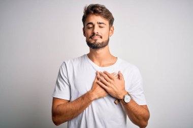 Sakallı, sıradan tişörtlü, beyaz arka planda duran, elleri göğsünde, gözleri kapalı ve yüzünde minnet ifadesi olan genç yakışıklı bir adam. Sağlık kavramı.