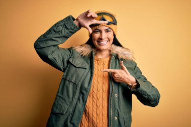 Kış havası için kayak gözlüğü ve palto takan genç ve güzel bir İspanyol kadın gülümsüyor ve elleri ve parmakları gülen yüzüyle çerçeve yapıyor. Yaratıcılık ve fotoğrafçılık kavramı.