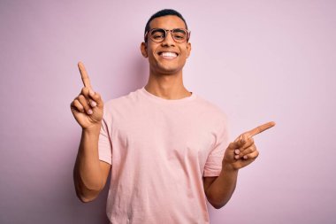 Yakışıklı Afro-Amerikan erkeği pembe arka planda günlük tişört ve gözlük takıyor. Farklı yönleri işaret ederek gülümsüyor. Reklam için boşluk kopyala