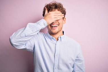 Sarışın, kıvırcık saçlı, beyaz arka planda çizgili tişört giyen, gülümseyen ve elleri yüzünde, şaşırtmak için gözleri kapatan genç bir adam. Kör kavram.