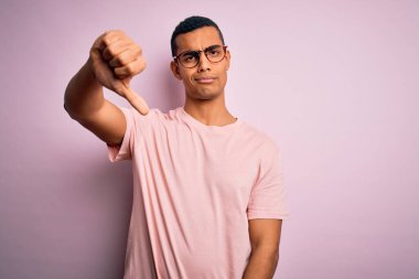 Yakışıklı Afro-Amerikalı adam pembe arka planda günlük tişört ve gözlük takıyor mutsuz, kızgın, reddedilmiş ve olumsuz jestler yapıyor. Kötü ifade.