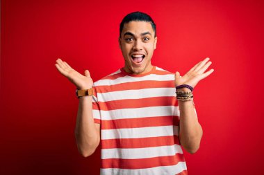 İzole edilmiş kırmızı arka planda dikilmiş sıradan çizgili tişört giyen genç Brezilyalı adam çılgın ve başarıyı kollarını kaldırmış ve gözleri açık bir şekilde heyecanla haykırarak kutluyor. Kazanan konsept