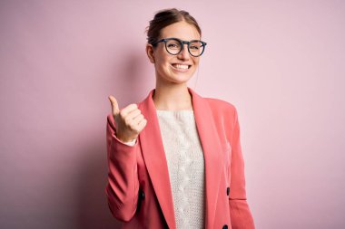 Ceket ve gözlük takan genç, güzel bir kızıl kadın pembe arka planda mutlu bir el hareketi yapıyor. Kameraya bakıp başarıyı gösteren ifade onaylanıyor.
