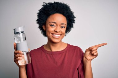 Kıvırcık saçlı, genç Afrikalı Afro kadın ferahlatıcı olarak su içiyor. Elini ve parmağını kenarı göstererek mutlu bir şekilde işaret ediyor.