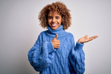 Beyaz arka plan üzerine balıkçı yaka kazak giyen genç ve güzel Afrikalı Amerikalı kadın avuç içini gösteriyor ve baş parmaklarıyla iyi hareket ediyor, mutlu ve neşeli gülümsüyor.