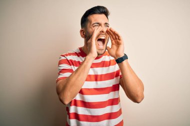Sıradan çizgili tişört giyen yakışıklı genç adam izole edilmiş beyaz arka planda duruyor ve elleri ağzında kızgın bir şekilde bağırıyor.
