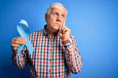 Üst düzey yakışıklı, elinde mavi kanser kurdelesi olan bir adam izole edilmiş arka planda ciddi bir yüz soru soran, kafası karışmış bir fikir.