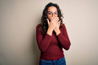 Kıvırcık saçlı, günlük süveter giyen ve beyaz arka planda gözlük takan güzel bir kadın ağzını yanlışlıkla ellerle kapatarak şok etti. Gizli konsept.
