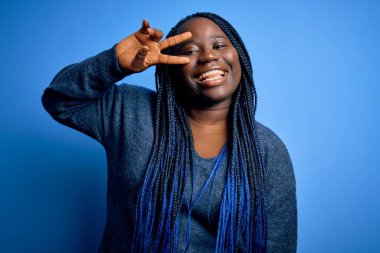 Mavi arka planda günlük süveter giyen Afrikalı Amerikalı artı cüsseli kadın. Yüzünde parmaklarıyla barış sembolü yapıyor. Gülümsüyor, zaferini gösteriyor.