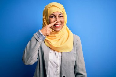 Orta yaşlı esmer iş kadını, geleneksel Müslüman tesettüre bürünmüş mavi arka planda el ve burnu işaret eden, neşeli gülümseyen. Güzellik konsepti