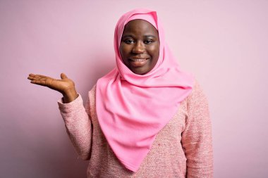 Genç Afro-Amerikan artı cüsseli kadın izole edilmiş pembe arka plan üzerine Müslüman başörtüsü takıyor neşeli bir şekilde sunum yapıyor ve kameraya bakıyor..