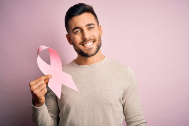 Genç ve yakışıklı bir adam pembe kanser kurdelesi taşıyor. İzole edilmiş arka planda destek sembolü. Mutlu bir yüzle ayakta duruyor ve gülümsüyor.