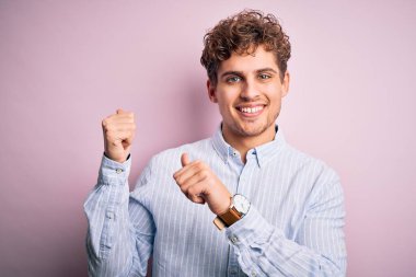 Sarışın, kıvırcık saçlı, beyaz arka planda çizgili tişört giyen, eli ve başparmağı havada, kendine güvenen genç bir adam.