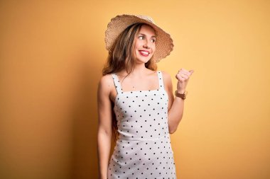 Sarı arka planda yaz şapkası takmış, gülümseyen, gülümseyen ve baş parmağıyla yüzünü gösteren genç, güzel sarışın kadın..