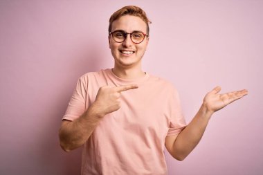 İzole edilmiş pembe arka plan üzerinde duran gündelik tişört giyen genç ve yakışıklı adam kameraya hayret ediyor ve gülümsüyor..